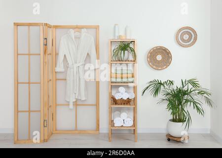 Weiche gefaltete Handtücher auf Holzregal und Bademantel nahe der weißen Wand Stockfoto