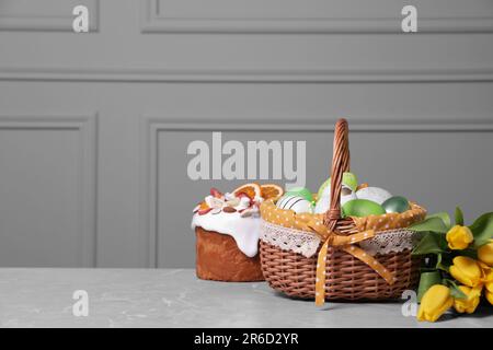 Osterkorb mit vielen bemalten Eiern in der Nähe von leckerem Kuchen und einem Strauß Tulpen auf einem grau strukturierten Tisch. Platz für Text Stockfoto