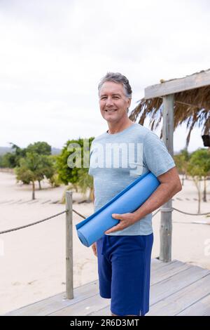 Porträt eines lächelnden weißen Seniorenmannes mit blauer Trainingsmatte im Touristenresort Stockfoto