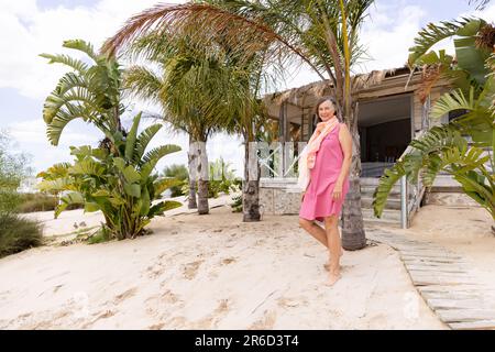 Eine weiße Seniorin, die am Sandstrand gegen Palmen und Hütten spaziert Stockfoto