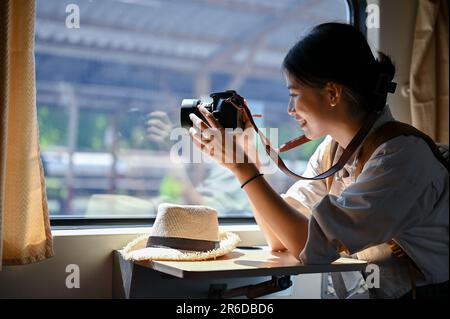 Eine glückliche und wunderschöne junge asiatische Reisende setzt sich an ihren Platz und macht mit ihrer Kamera ein Foto vom Zug. Sommerurlaub, Reise, Solo Stockfoto