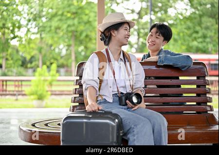 Ein hübsches, junges asiatisches Touristenpaar mit ihren Koffern spricht gerne auf einer Bank auf dem Bahnsteig, bevor es gemeinsam in den Zug steigt. Stockfoto