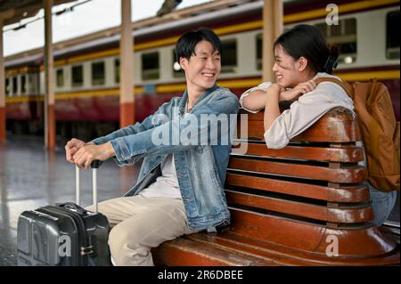 Ein hübsches, junges asiatisches Touristenpaar mit ihren Koffern spricht gerne auf einer Bank auf dem Bahnsteig, bevor es gemeinsam in den Zug steigt. Flitterwochen, Stockfoto