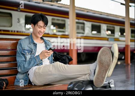 Ein gutaussehender und glücklicher junger männlicher asiatischer Rucksacktourist oder Reisender, der sein Smartphone auf einer Bank benutzt, während er auf seinen Zug am Bahnsteig wartet. Rucksacktouristen, Stockfoto
