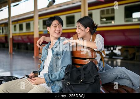 Ein hübsches, junges asiatisches Touristenpaar spricht gerne auf einer Bank auf dem Bahnsteig, bevor es gemeinsam in den Zug steigt. Flitterwochen, Touristenpaar, Trav Stockfoto