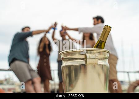 Nahaufnahme einer Flasche Sekt in einem Eiskübel, während eine vielfältige Gruppe von Freunden fröhlich im Hintergrund anstoßen. Die Freunde, unscharf, C. Stockfoto