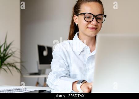 Junge brünette Geschäftsfrau in Brille, die lächelt und im Büro an einem Laptop arbeitet Stockfoto