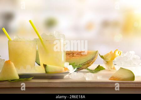 Zwei Gläser mit Eismelonengetränk auf einem Holztisch voller zerdrücktem Eis und geschnittenem Obst mit Küche im Hintergrund. Vorderansicht. Waagerecht c Stockfoto