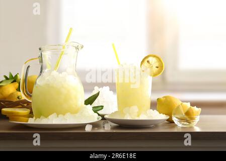Limonade mit Eis in einem Krug und Glas auf einer Holzbank mit Obst und zerstoßenem Eis mit einem sonnigen Fenster im Hintergrund in einer Küche. Vorderansicht. Stockfoto