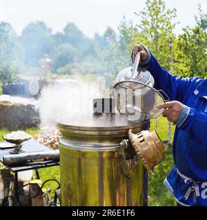 Ein Mann gießt Wasser aus einem Wasserkocher in einen Samovar durch ein Sieb, einen alten russischen Samovar mit Holzfeuer. Stockfoto