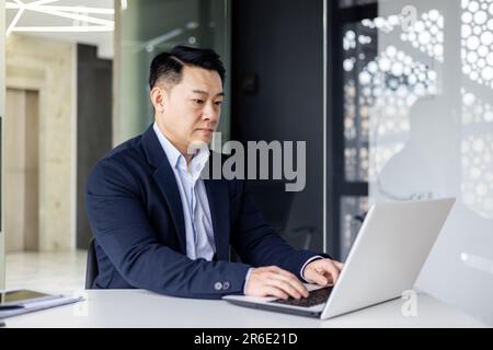 Seriöser und umsichtiger Geschäftsmann, der im Büro arbeitet, am Tisch sitzt und ein Notebook benutzt, ein erfahrener asiatischer Boss denkt und tippt über die Tastatur. Stockfoto