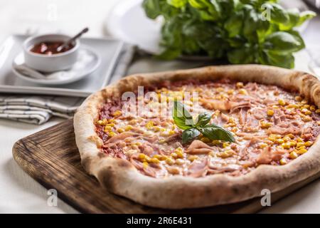 Italienische Pizza mit Mais und Basilikumblättern, serviert auf einem Holzbrett. Stockfoto