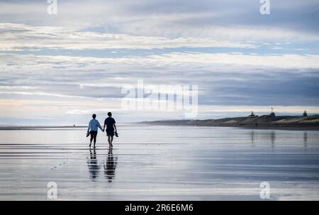 Ein Paar, das Händchen hält, Schuhe hält und barfuß am Muriwai Beach spaziert. Auckland. Stockfoto