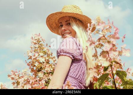 Lächelnde junge blonde Frau mit Strohhut in der Nähe von Hortensien-Blumen Stockfoto