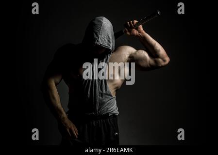 Gefährlicher muskulöser Mann in der Maske mit Baseballschläger bereit zum Kampf. Ein aggressiver Kapuzenmann hält einen Baseballschläger auf Schwarz. Stockfoto