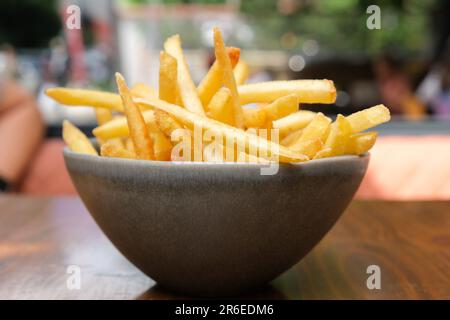 Aus nächster Nähe gebratene Kartoffeln in der Schüssel auf einem Holztisch mit isoliertem Restaurant im Hintergrund. Selektiver Fokusbereich. Stockfoto