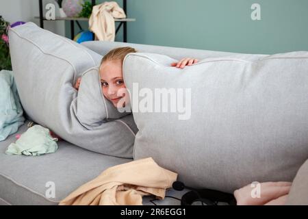 Ein Mädchen spielt Verstecken. Das Kind hat Dinge im Zimmer verstreut. Sie will ihre Kleider nicht wegräumen. Stockfoto