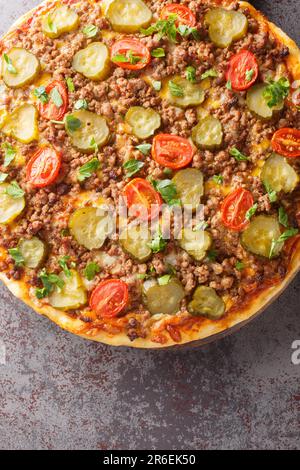 Köstliche heiße Hamburger-Pizza mit Rinderhack, Tomaten, Gurken und Käse aus nächster Nähe auf einem Holzbrett auf dem Tisch. Vertikale Draufsicht von oben Stockfoto