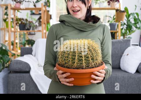 Großer echinocactus Gruzoni in den Händen einer überraschten und lustigen Emotionen Frau im Inneren eines grünen Hauses mit Regal-Sammlungen von Hauspflanzen. Hom Stockfoto