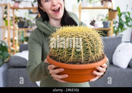 Großer echinocactus Gruzoni in den Händen einer überraschten und lustigen Emotionen Frau im Inneren eines grünen Hauses mit Regal-Sammlungen von Hauspflanzen. Hom Stockfoto