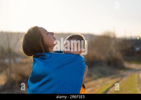 Eine Frau umarmt ihren kleinen Sohn, eingewickelt in die gelb-blaue Flagge der Ukraine im Freien. Stockfoto