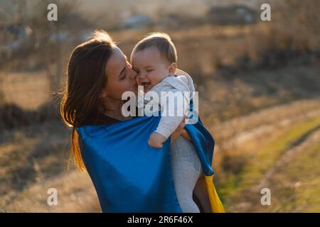 Eine Frau umarmt ihren kleinen Sohn, eingewickelt in die gelb-blaue Flagge der Ukraine im Freien. Stockfoto