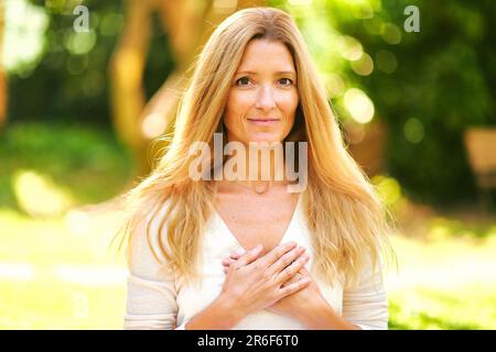 Außenporträt einer schönen Frau, die sich im sonnigen Park oder Garten ruht, Hände an der Brust hält, Dankeschön Stockfoto