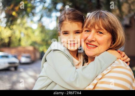 Ein kleines Mädchen, das eine lächelnde Frau mittleren Alters umarmt. Das süße weibliche Kind und ihre Großmutter gehen gern draußen spazieren. Oma und ihre Enkelin haben tolle Tage Stockfoto