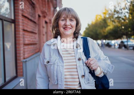 Attraktive blonde Studentin mittleren Alters mit lockigem Haar in stilvoller Jeanskleidung, die in der Stadt spaziert. Bildungs- und Studienkonzept Stockfoto