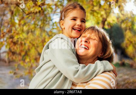 Ein kleines Mädchen, das eine lächelnde Frau mittleren Alters umarmt. Das süße weibliche Kind und ihre Großmutter gehen gern draußen spazieren. Oma und ihre Enkelin haben tolle Tage Stockfoto