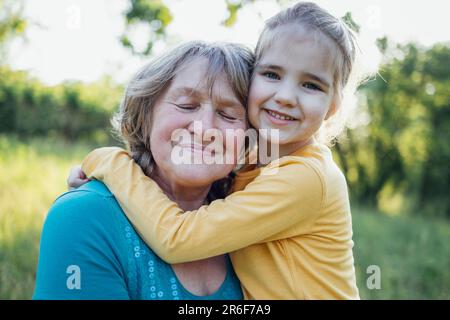 Ein kleines Mädchen, das eine lächelnde Frau mittleren Alters umarmt. Das süße weibliche Kind und ihre Großmutter gehen gern draußen spazieren. Oma und ihre Enkelin haben tolle Tage Stockfoto