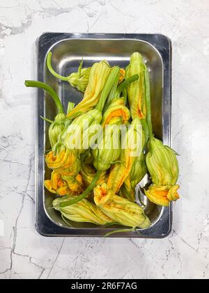 Nahaufnahme von Zucchini-Blumen, die auf einem Metalltablett auf einem Marmortisch liegen. Gemüse kochen. Heller, farbiger Naturhintergrund. Speicherplatz kopieren. Stockfoto