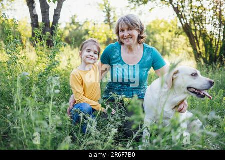 Die grauhaarige Großmutter und die süße kleine Enkelin gehen mit ihren Hunden im Park spazieren. Ältere wunderschöne Frau streichelt goldenes labrador und Stockfoto