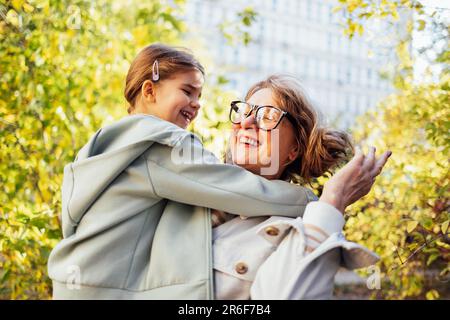 Ein kleines Mädchen, das eine lächelnde Frau mittleren Alters umarmt. Das süße weibliche Kind und ihre Großmutter gehen gern draußen spazieren. Oma und ihre Enkelin haben tolle Tage Stockfoto