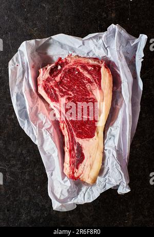 Prime Bone-in-Ribeye-Steak in Metzgerpapier verpackt auf schwarzem Marmorhintergrund. Stockfoto