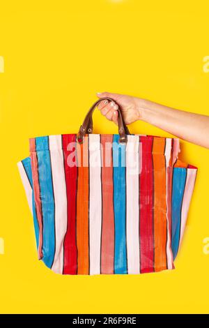 Frauenhand mit einer bunten gestreiften Plastik-Einkaufstasche auf gelbem Hintergrund Stockfoto