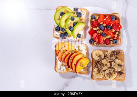 Beerensandwiches. Verschiedene Toasts mit Erdnussbutter, Frischkäse, Schokoladenaufstrich mit Sommerbeeren und Obst - Erdbeeren, Bananen, Pfirsich, Stockfoto