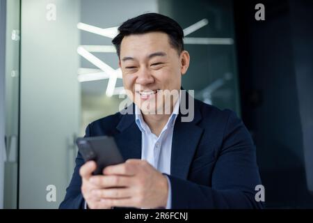 Asiatischer Geschäftsmann im Büro, der telefoniert, hält der Chef ein Smartphone in der Hand, liest Nachrichten und schreibt lächelnd eine Nachricht. Stockfoto