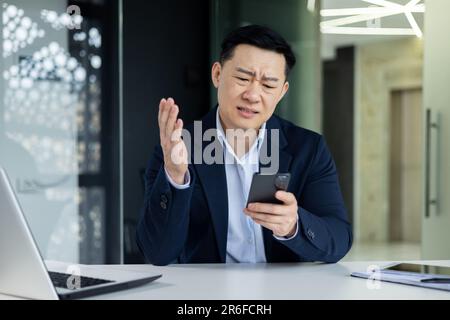 Aufgewühlter asiatischer Mann im Büro, der schlechte Nachrichten vom Telefon im Internet liest, Geschäftsmann in der Nähe des Fensters, der im Büro arbeitet, mit Smartphone Stockfoto