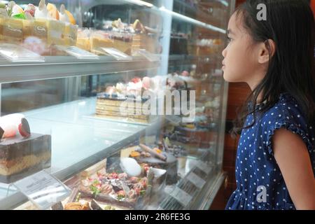 Asiatisches kleines Mädchen, das Kuchen und Süßigkeiten in einer Bäckerei ansieht. Stockfoto