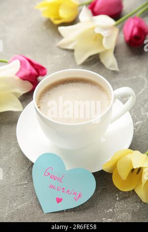 Kaffeebecher mit Tulpenblumen und Noten Guten Morgen auf grauem Hintergrund, Frühstück am Muttertag oder Frauentag. Vertikales Foto. Draufsicht Stockfoto