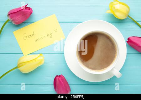 Kaffeebecher mit Tulpenblumen und Noten Guten Morgen auf blauem Hintergrund, Frühstück am Muttertag oder Frauentag. Draufsicht Stockfoto