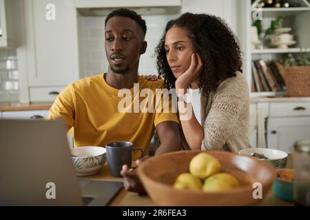 Ein junges, multiethnisches Paar, das mit einem Laptop online surft, während es morgens zusammen in der Küche frühstückt Stockfoto