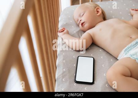 Kleines Baby in der Windel schläft sicher im Bett mit Telefon und spielt Musik im Schlafzimmer, während die Eltern sich im Wohnzimmer ausruhen. Frohe Mutterschaft und Technologie. Smartphone-Modell für die Anwendung zum Schlafen von Babys Stockfoto