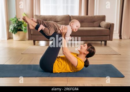 Neue mutter hebt ihr niedliches kleines Baby mit den Beinen, macht postnatales Fitnesstraining. Sportliche Mutter ist mit einem Baby zu Hause in Fitness und Yoga beschäftigt. Gesunde Mutterschaft und postpartale Genesung Stockfoto