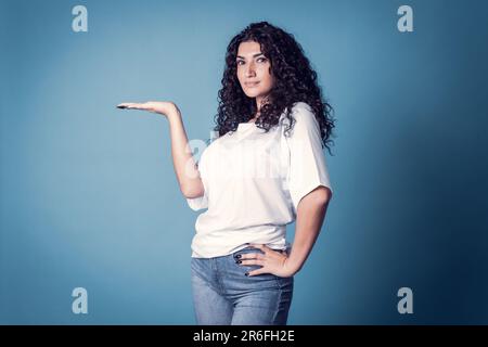 Aufnahme einer selbstbewussten, geschwungenen Frau, trägt ein legeres, einfarbiges weißes T-Shirt, das isoliert auf blauem Hintergrund gehalten wird. Stockfoto