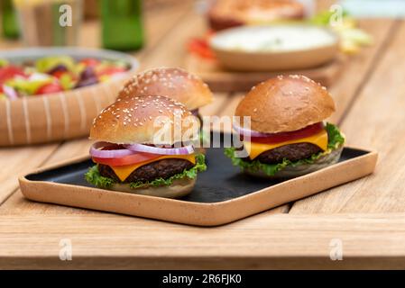 Drei vegane Burger auf einem Holzteller mit verschiedenen veganen Tellern auf einem Holztisch Stockfoto