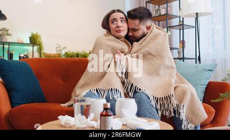 Junge kranke Familie verheiratete Frau, die an Kälte oder Allergie leidet, auf dem Heimsofa im Wohnzimmer. Die kranke Ehefrau bläst Niesen, Rotz in die Serviette. Coronavirus-Quarantäne, Pandemie Stockfoto