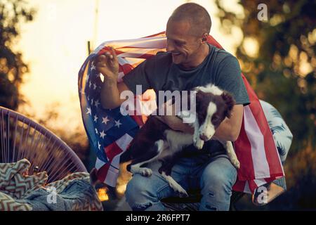Ein Mann mittleren Alters mit der US-Flagge auf dem Rücken kuschelt mit dem Hündchen Border Collie, draußen in einem gemütlichen Stuhl, bei Sonnenuntergang. 4. Juli, Unabhängigkeitstag Stockfoto