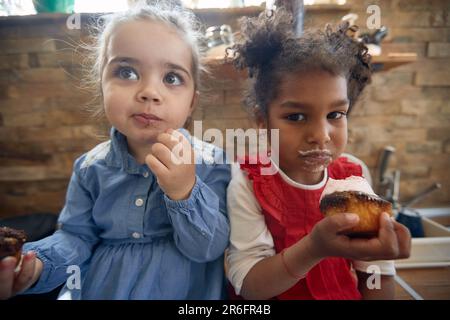 Nahaufnahme von zwei hübschen kleinen Mädchen, die Muffins essen und süß aussehen. Zuhause, Familie, Lifestyle-Konzept. Stockfoto